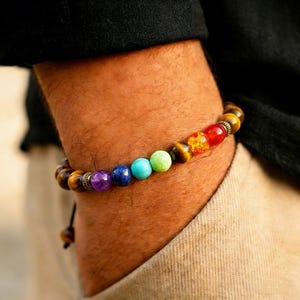 May include: A beaded bracelet featuring a variety of colorful gemstones, including purple, blue, turquoise, green, yellow, orange, and red. The bracelet is strung on a black cord and has a tiger eye stone bead.