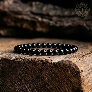 May include: A black beaded bracelet composed of polished, round beads. The bracelet is displayed on a rustic, wooden surface, with a dark, blurred background. The beads appear to be approximately 0.3 inches in diameter.