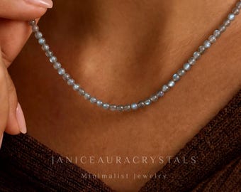 May include: A delicate necklace composed of small, round, light blue beads. The necklace is displayed on a person's neck, with a hand gently holding the chain. The text "JANICEAURACRYSTALS Minimalist Jewelry" is visible at the bottom.