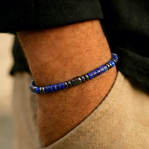 Puede incluir: Una pulsera de cuentas con pequeñas cuentas cuadradas de color azul oscuro, intercaladas con cuentas de color gris oscuro y negro. La pulsera tiene una cuenta central hexagonal negra. La pulsera se lleva en la muñeca.