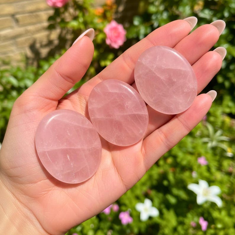 May include: Three smooth, oval-shaped rose quartz stones. The stones are a soft pink color and appear polished. The stones are held in a hand, with a blurred background of green foliage and pink flowers.