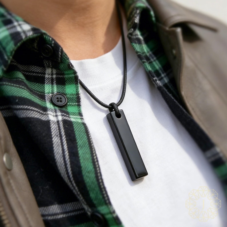 May include: A black rectangular pendant necklace. The pendant is suspended from a black cord and hangs against a white t-shirt. The person is wearing a green and black plaid shirt and a brown leather jacket.