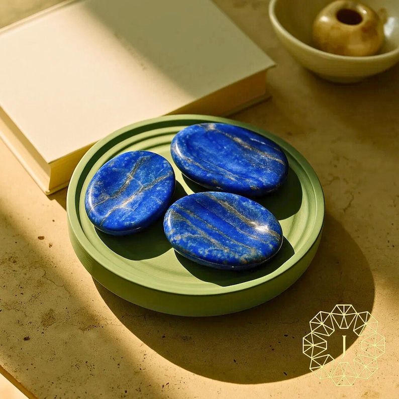 May include: Three oval-shaped lapis lazuli stones with gold veining rest on a green ceramic tray. The stones are a deep blue color. A book and a small bowl are in the background.