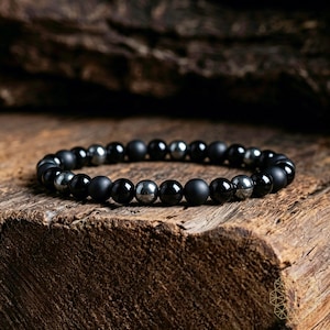 Könnte beinhalten: Ein schwarzes Perlenarmband mit einer Mischung aus matten und polierten runden Perlen. Das Armband enthält eine Kombination aus schwarzen und metallisch-silbernen Perlen, die auf einem elastischen Band aufgereiht sind. Das Armband wird auf einer Holzoberfläche präsentiert.