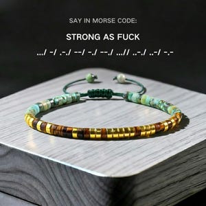 May include: A beaded bracelet with a mix of turquoise, brown, and gold-colored beads. The bracelet has a sliding knot closure and the text "STRONG AS FUCK" in Morse code.