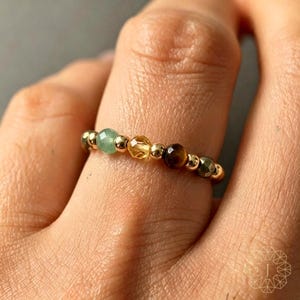 Könnte beinhalten: Ein Perlenring bestehend aus einer Abfolge kleiner goldfarbener Metallkugeln, die mit facettierten Edelsteinen in den Farben Hellgrün, Gelb, Braun und Dunkelgrau durchsetzt sind und einen kreisförmigen Ring am Finger bilden.