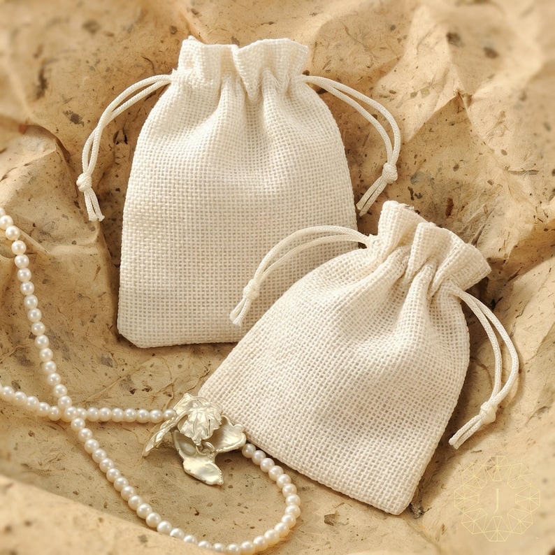 May include: Two small, off-white drawstring pouches made of a woven fabric with a visible grid texture. The pouches feature matching off-white cords for closure. A long strand of white pearls with a metallic leaf-shaped pendant is draped nearby.