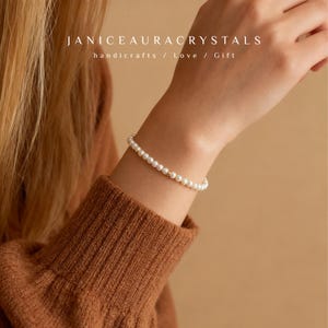 May include: A delicate pearl bracelet with small, round white pearls and gold accents. The bracelet is worn on a wrist, and the image includes the text "JANICEAURACRYSTALS handicrafts / Love / Gift". The bracelet is a piece of jewelry.