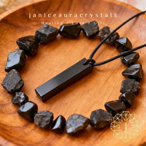 May include: A black rectangular pendant necklace with a black cord, surrounded by a necklace of dark, rough-cut stones. The image includes the text "janiceauracrystals Healing / Love / Gift" in white.
