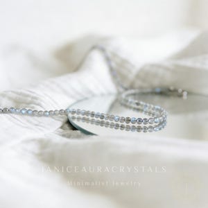May include: A delicate necklace made of small, round, gray and blue beads. The necklace is displayed on a reflective surface, with the text "JANICEAURACRYSTALS Minimalist Jewelry" in the bottom center.