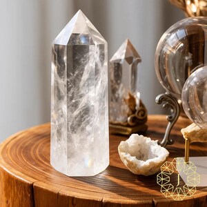 May include: A clear quartz crystal point stands on a wooden table, showcasing its multifaceted design. The crystal is approximately 15 cm tall. Other crystals and decorative objects are in the background, creating a serene and elegant display.