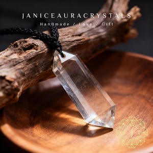 May include: A clear quartz crystal pendant with a black cord. The crystal is a long, hexagonal shape. The text "JANICEAURACRYSTALS Handmade / Love / Gift" is at the top of the image. The pendant is displayed on a wooden surface.