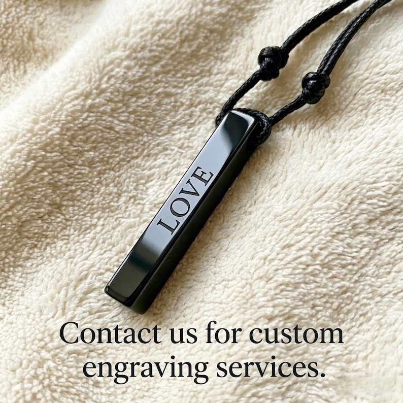 May include: A black rectangular pendant necklace with the word "LOVE" engraved in white. The pendant hangs from a black cord with adjustable knots. The background is a soft, textured, off-white fabric.