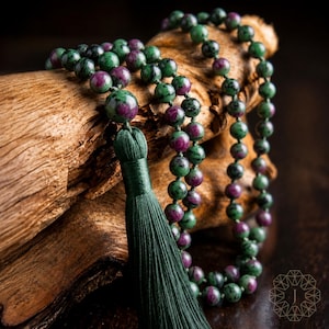 May include: A mala necklace featuring round beads of green and magenta stone, strung together with knots. The necklace includes a long, dark green silk tassel attached to the main bead. The beads display a mottled pattern of dark green and deep pink.