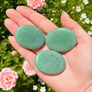 May include: Three smooth, oval-shaped green aventurine stones are held in a hand. The stones are a soft, muted green color. The text "JANICEAURACRYSTALS Handcrafted jewelry" is visible at the top of the image.