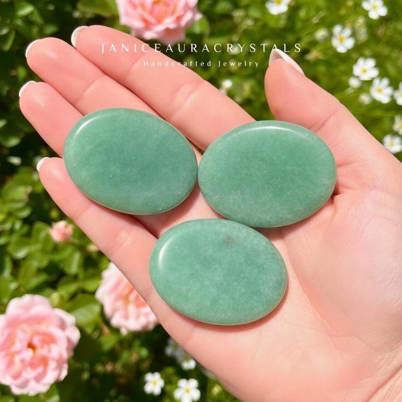 May include: Three smooth, oval-shaped green aventurine stones are held in a hand. The stones are a soft, muted green color. The text "JANICEAURACRYSTALS Handcrafted jewelry" is visible at the top of the image.