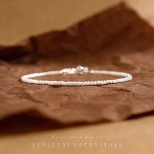 Könnte beinhalten: Ein zartes, weißes Perlenarmband mit Silberverschluss, präsentiert auf einer braunen, strukturierten Oberfläche. Das Armband besteht aus kleinen, runden Perlen. Der Text "Handcrafted jewelry" und "JANICEAURACRYSTALS" ist sichtbar.