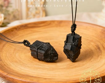 Peut inclure: Deux pendentifs en cristal de tourmaline noire sur une assiette en bois. Chaque cristal est enveloppé de fil noir et suspendu à un cordon noir. Le texte "janiceauracrystals Handmade / Love / Gift" est visible en haut.