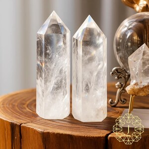 May include: Two clear quartz crystal points with a hexagonal base and a pointed top. The crystals are displayed on a wooden surface. The crystals are transparent with internal inclusions. The crystals are part of a collection of crystals and decorative objects.