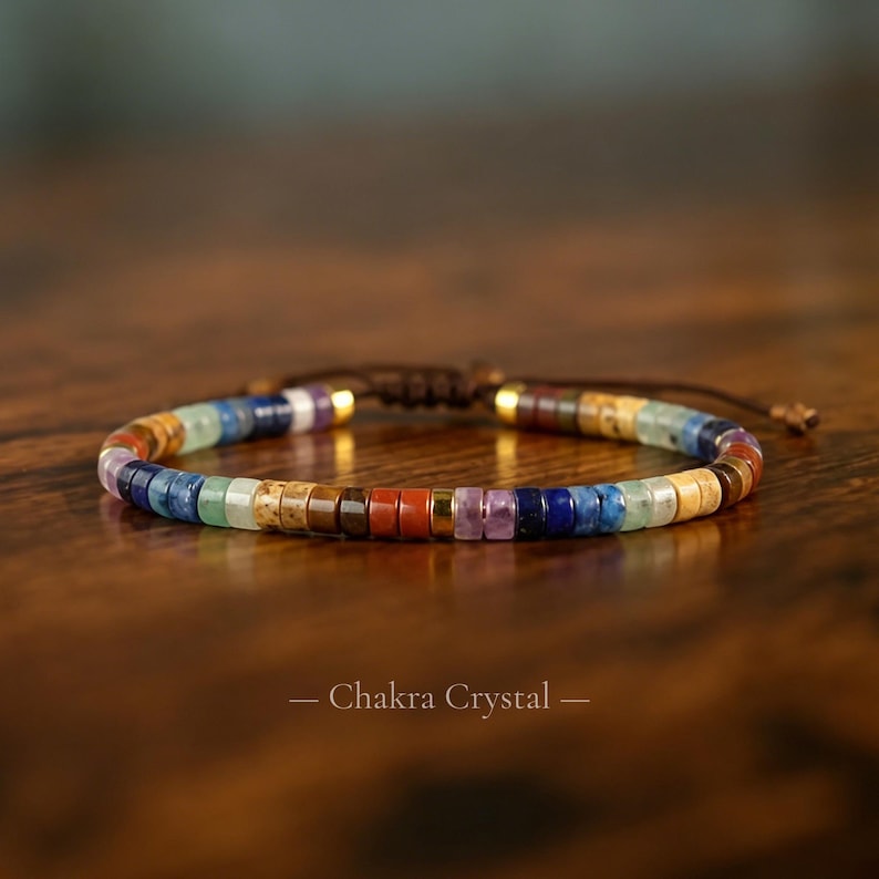 May include: A bracelet featuring a sequence of small, flat, circular beads in various colors including red, orange, yellow, green, blue, purple, and brown, interspersed with metallic gold spacers and finished with a dark brown adjustable cord. The text Chakra Crystal is visible.