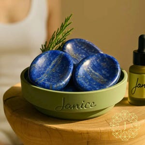 May include: A collection of deep blue lapis lazuli stones in a green bowl with the name "Janice" inscribed. The stones have gold flecks and are displayed with a sprig of greenery. A small bottle with a dropper is visible.