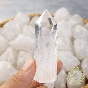 May include: A clear quartz crystal point with a faceted tip, held in a hand. The crystal is surrounded by other white quartz stones in a woven basket. The crystal is approximately 10 cm tall.