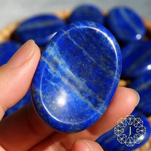 May include: A polished, oval-shaped lapis lazuli stone, held in a hand, showcasing a deep blue color with white and gold veining. Other similar stones are visible in the background, suggesting a collection of gemstones.