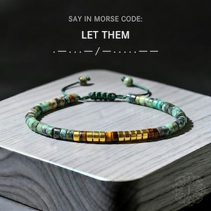 May include: Adjustable beaded bracelet with turquoise and gold-colored beads. The bracelet features a central section of gold-colored beads and a green cord. The text "SAY IN MORSE CODE: LET THEM" is at the top.