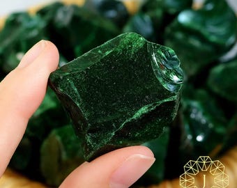 Puede incluir: Una mano sostiene una piedra verde oscuro de textura rugosa, mostrando su forma natural e irregular. La piedra tiene una superficie brillante con estructuras internas visibles. Otras piedras similares están en una cesta de mimbre en el fondo. La imagen resalta el color y la textura de la piedra.