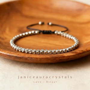 May include: A silver beaded bracelet with a black adjustable cord, resting on a wooden bowl. The bracelet features small, cube-shaped beads. The text "janiceauracrystals" is visible, along with "Love • Ritual".