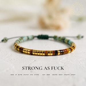 Peut inclure: Un bracelet composé de perles en forme de disque, alternant entre le doré métallique, l'œil de tigre brun et le turquoise. Il est doté d'un cordon de serrage vert foncé et porte l'inscription STRONG AS FUCK avec des points et des tirets morse.