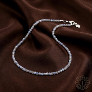 May include: A delicate necklace composed of small, round, iridescent beads. The beads are primarily light gray with subtle blue and white highlights. The necklace has a silver clasp with a small heart charm. The necklace rests on a dark brown, textured fabric.