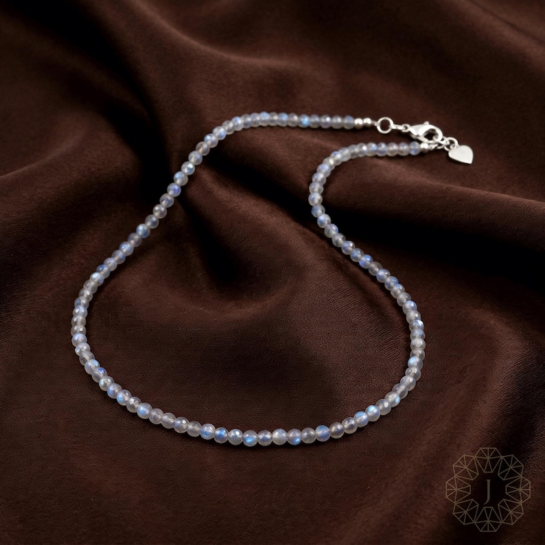 May include: A delicate necklace composed of small, round, iridescent beads. The beads are primarily light gray with subtle blue and white highlights. The necklace has a silver clasp with a small heart charm. The necklace rests on a dark brown, textured fabric.
