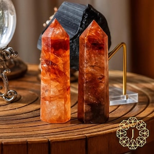 May include: Two orange and brown crystal towers with pointed tops stand on a wooden surface. The crystals have a translucent appearance, with visible internal patterns. A black stone and a decorative gold stand are in the background.