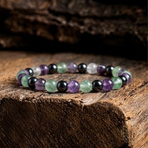 May include: A beaded bracelet featuring alternating round polished beads in shades of purple, translucent green, glossy black, and clear white. The beads are strung together in a repeating pattern to form a circular band of smooth, spherical stones.