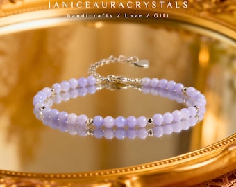 Puede incluir: Una delicada pulsera de cuentas lavanda con una cadena y cierre plateados. La pulsera se exhibe sobre una superficie reflectante, con el texto "JANICEAURACRYSTALS handicrafts / Love / Gift" visible en el fondo.