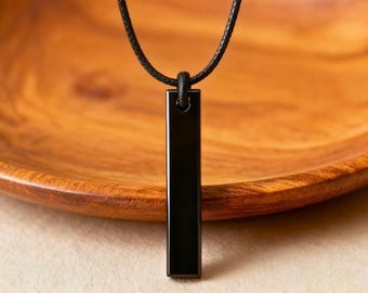 May include: A black rectangular pendant necklace hangs from a black cord. The pendant is a polished, solid black bar. The background is a wooden bowl with a natural wood grain.