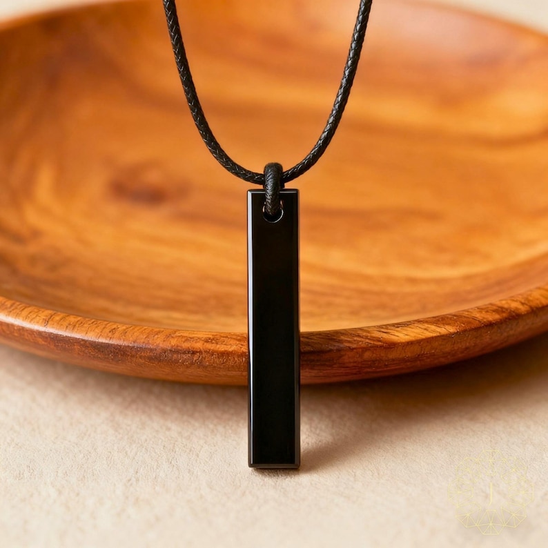 May include: A black rectangular pendant necklace hangs from a black cord. The pendant is a polished, solid black bar. The background is a wooden bowl with a natural wood grain.