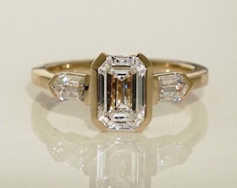 Puede incluir: Un anillo de oro con un diamante central de talla esmeralda, enmarcado por un bisel de oro, y dos diamantes baguette más pequeños y cónicos a cada lado. La banda del anillo es de oro liso.