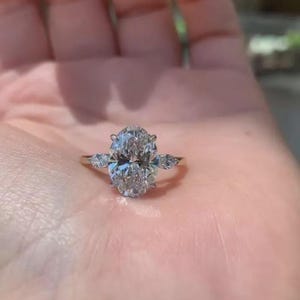May include: A gold ring featuring a large, oval-cut diamond as the centerpiece, flanked by two smaller pear-shaped diamonds. The ring's band is a polished gold, and the diamonds sparkle with multiple facets, reflecting light.