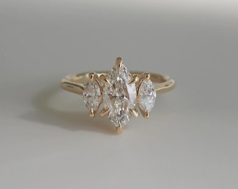 May include: A gold ring featuring a central marquise-cut diamond flanked by two smaller, pear-shaped diamonds. The ring's band is a simple, polished gold, and the diamonds sparkle brightly against a neutral background. This ring is a piece of fine jewelry.