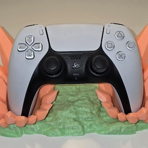 May include: A white and black PlayStation 5 controller resting on a 3D-printed stand. The stand features a light green base with peach-colored crystal-like formations on either side, creating a unique display for the gaming accessory.