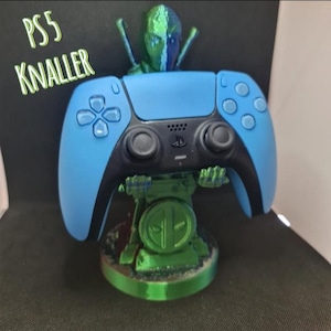 Könnte beinhalten: Ein blauer und schwarzer Gamecontroller, gehalten von einer grünen, irisierenden, 3D-gedruckten Figur mit kreisförmigem Sockel. Die Figur hat ein maskenartiges Gesicht und zwei Schwerter auf dem Rücken. Der Text PS5 KNALLER steht gelb links.