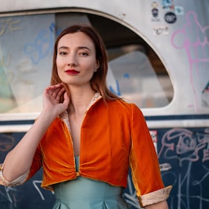 May include: A woman wearing a bright orange velvet cropped jacket with a blue and white patterned trim. The jacket is tied at the front with a gold ribbon. She is wearing a light blue dress underneath.