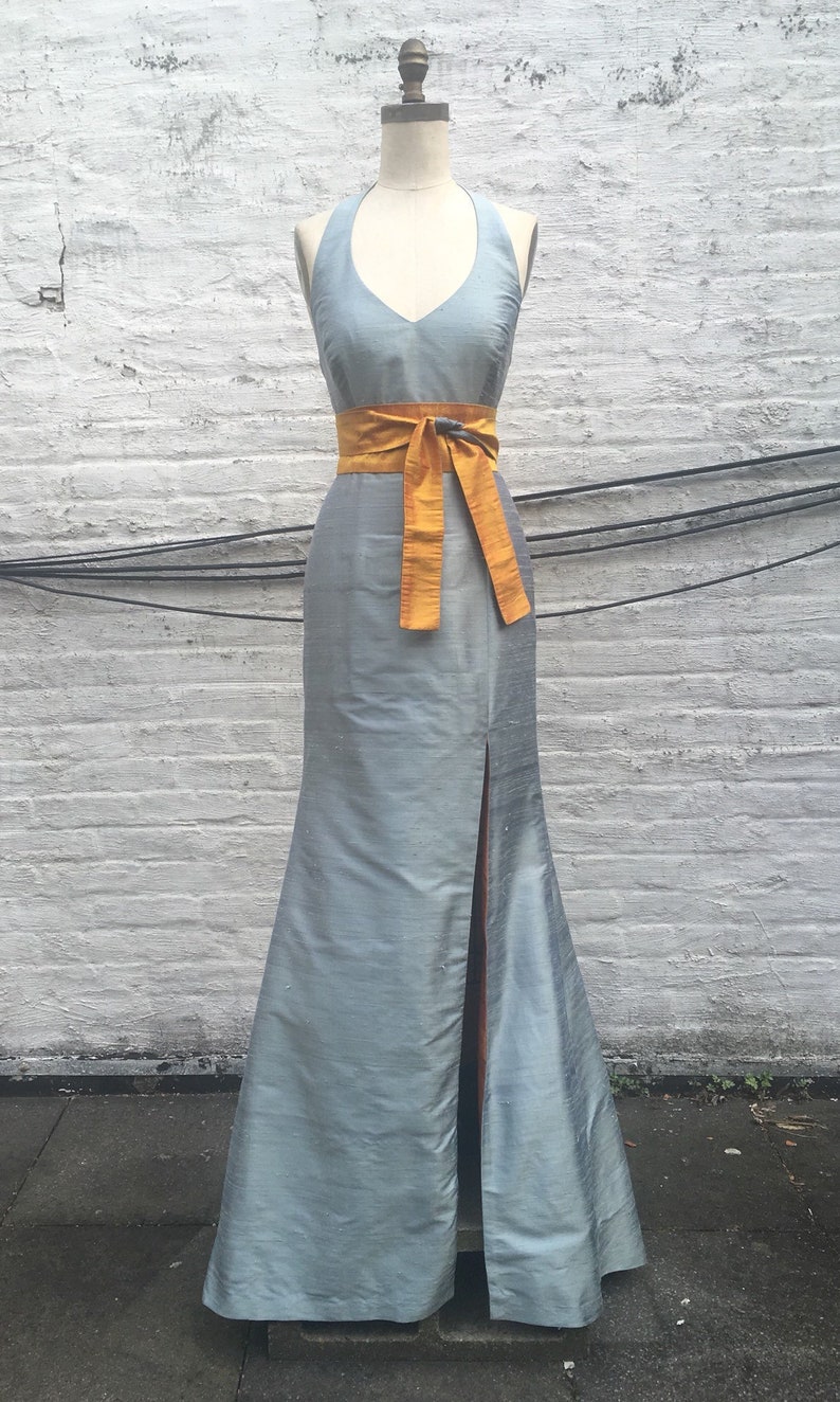 May include: A light blue halter neck gown with a floor-length skirt and a thigh-high slit. The dress features a mustard yellow sash tied at the waist. The gown is displayed on a mannequin against a white brick wall.