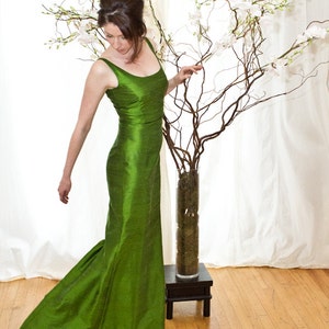 May include: A woman in a long, emerald green, silk dress stands in front of a white backdrop. She is looking down at a vase of white flowers.