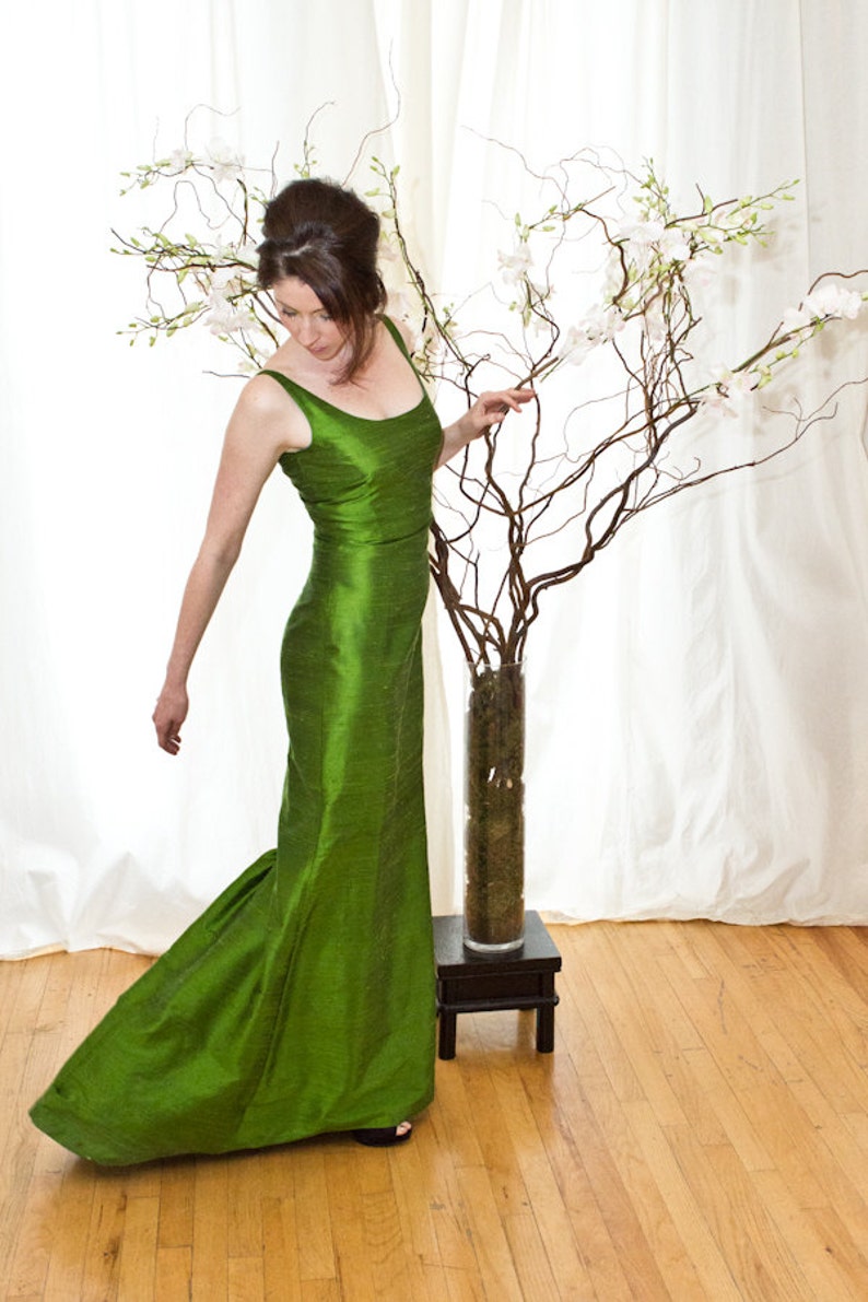 May include: A woman in a long, emerald green, silk dress stands in front of a white backdrop. She is looking down at a vase of white flowers.