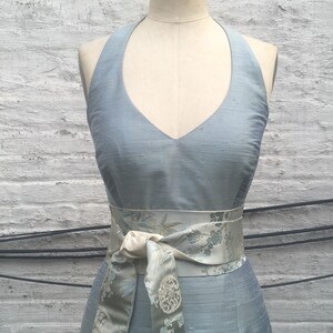 May include: A light blue halter dress on a mannequin. The dress features a V-neck and a matching sash with a floral pattern. The dress is made of a textured fabric, and the sash is tied in a bow at the waist.