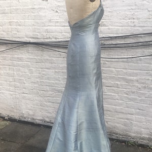 May include: A light blue, floor-length gown with a halter neckline and a mermaid silhouette. The dress is displayed on a mannequin against a white brick wall. The fabric has a subtle sheen.