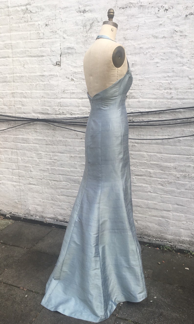 May include: A light blue, floor-length gown with a halter neckline and a mermaid silhouette. The dress is displayed on a mannequin against a white brick wall. The fabric has a subtle sheen.
